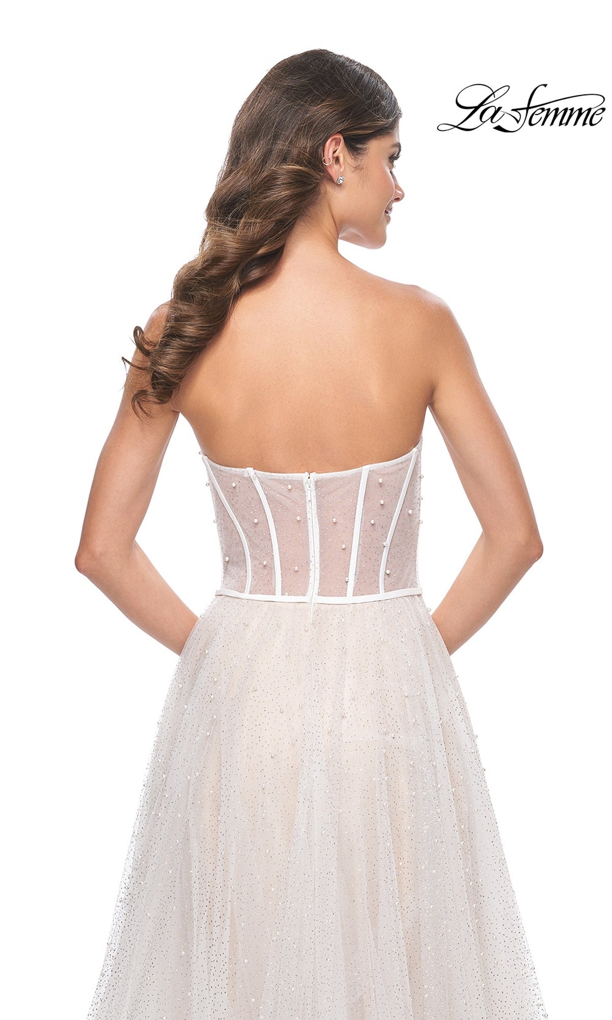 A woman with long, wavy hair is shown from the back wearing the La Femme Strapless Long White Prom Dress 32149, a pearl-embellished gown with a sheer corset bodice.